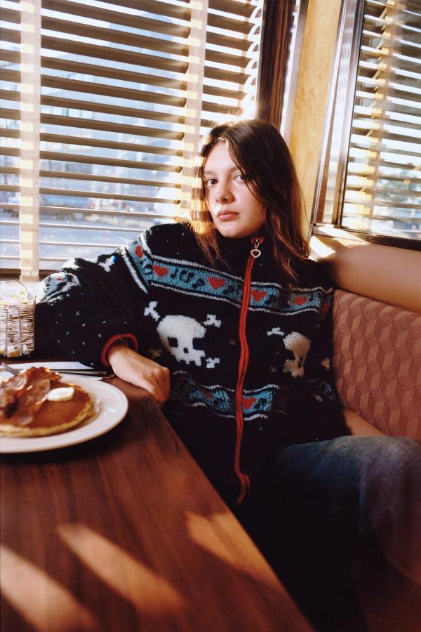 Model sits in a diner booth wearing a black multi sherpa jacket with red trim and a pattern of white skulls, hearts, and blue geometric designs.