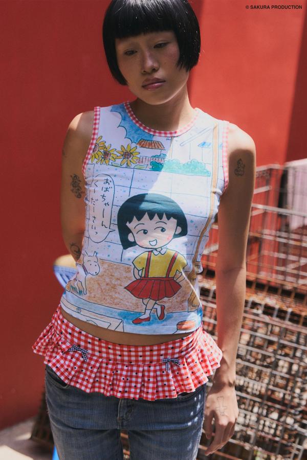 Woman wearing Sweet Shopper Tank. Top illustrates a manga-style image of a young girl and her dog walking into a store. She speaks in Japanese, "Please Grandma!" The top has a red gingham trim around the neck and sleeves. She wears jeans and a red gingham swim bottom over them against a red background with crates.