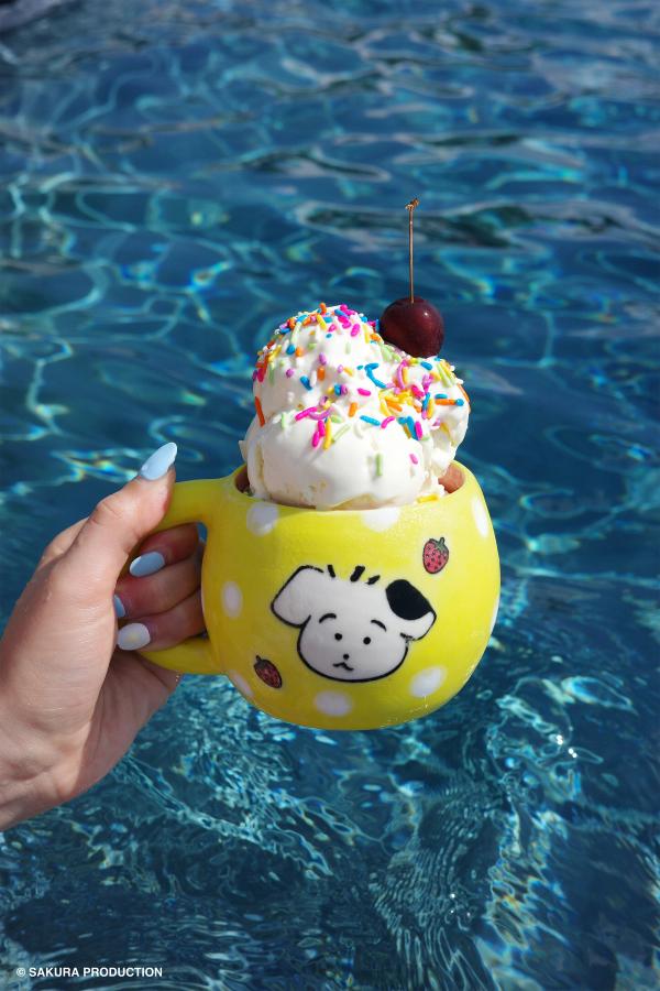 Chibi Maruko Chan hand made ceramic mug held against a pool backdrop. Yellow body and white polka dots with face of a little white and black dog surrounded by strawberries. There is ice cream with rainbow sprinkles and a cherry in the mug.