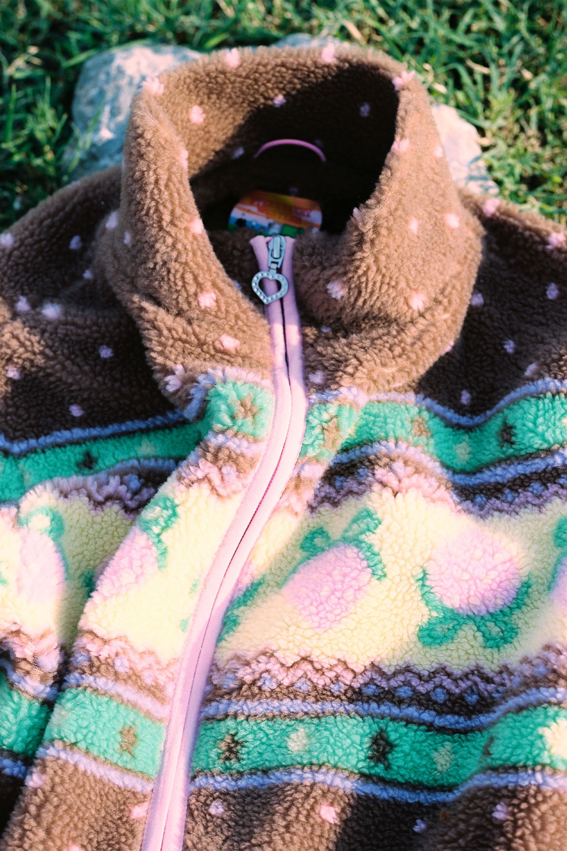 Fabric detail of the Brown fluffy jacquard knit sherpa jacket in a choco strawberry border motif with binding, zippers and a metal heart shape zipper pull.