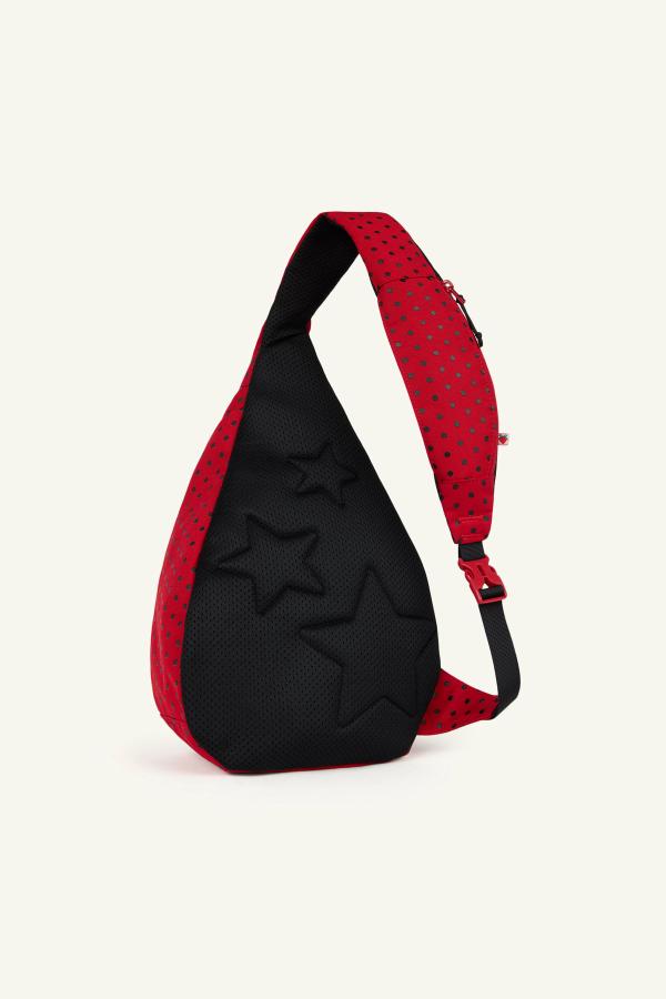 Product image of back view of a Red sling backpack with black polka dots and a black padded panel featuring embossed star shapes. The bag has a gray adjustable strap with a pink buckle and a small zip pocket on the strap.
