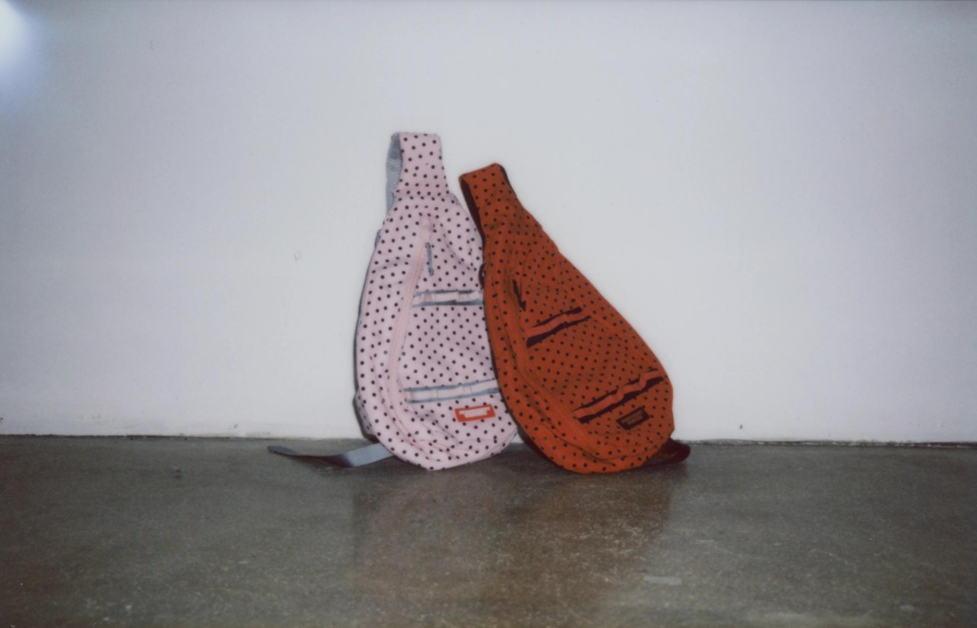 Polaroid photo of a pink Sling Bag and Red Sling Bag on the floor, leaning up against a white wall.