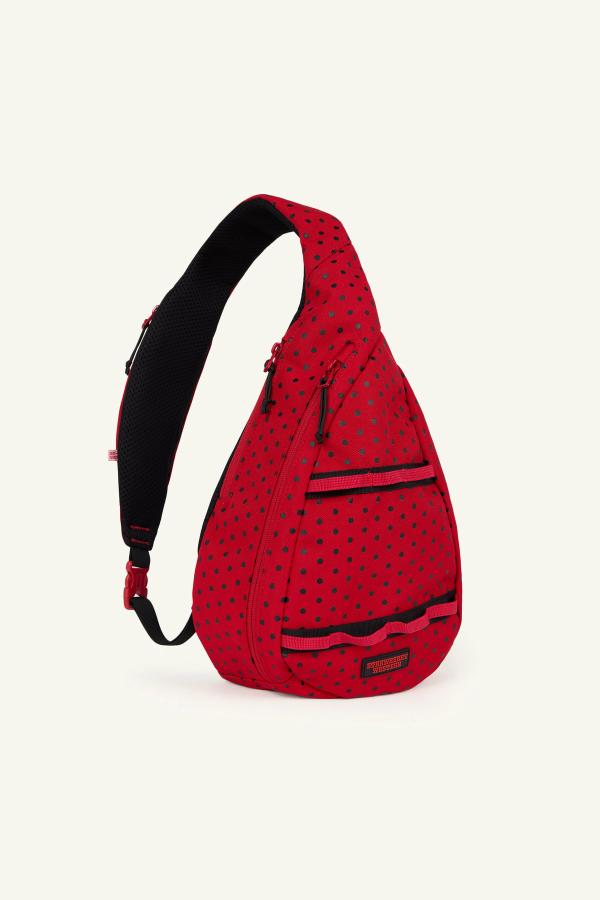Product image of front view of a Red sling backpack with black polka dots, gray zippers, and a black adjustable strap. The bag has multiple front zip compartments.