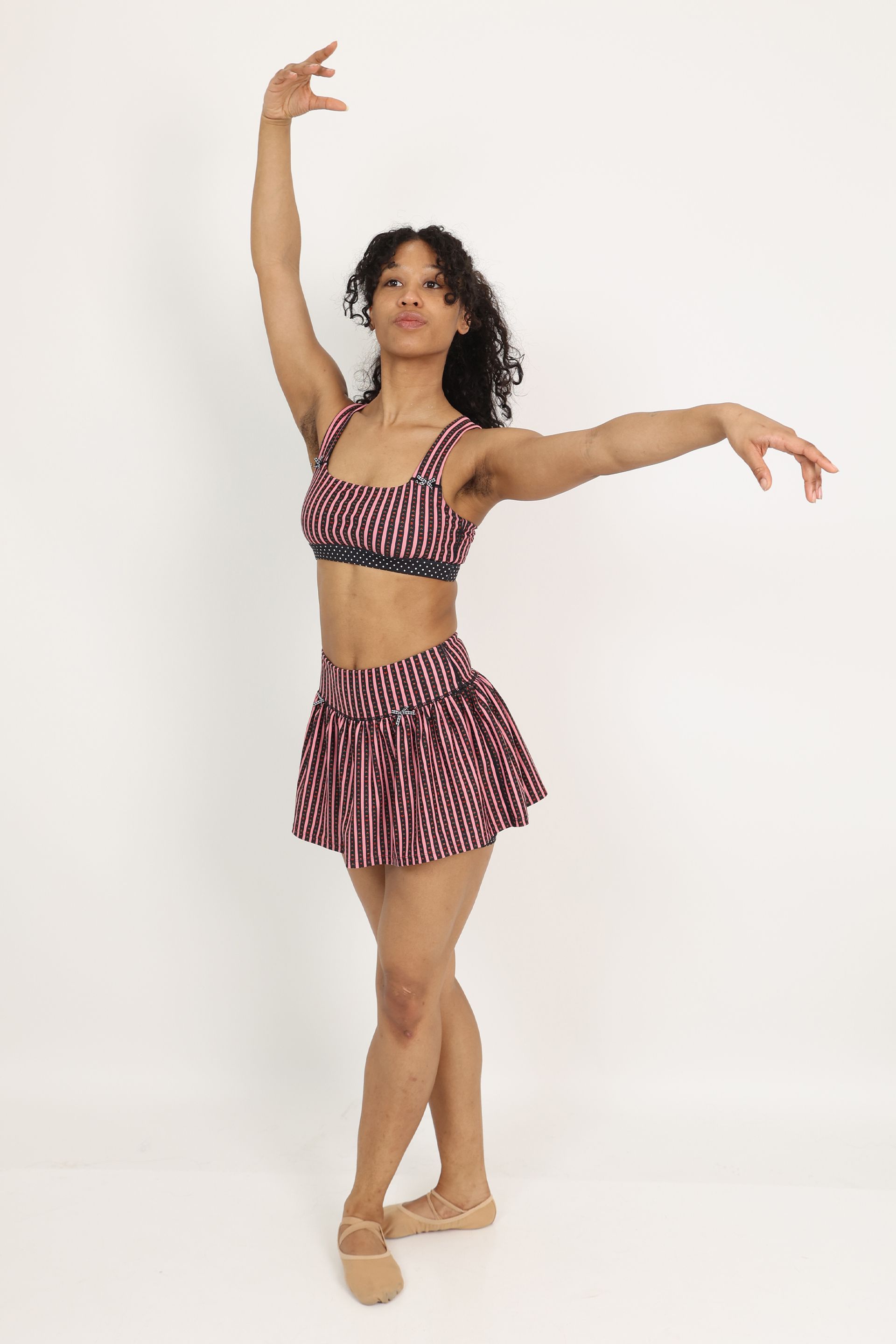 Model in a dance pose wearing the Coco Sports Bra and Rika Ruffle Skort featuring a hand-drawn, floral border stripe print  in Pink/Black with contrast polka dot print elastic band and gingham bow details.