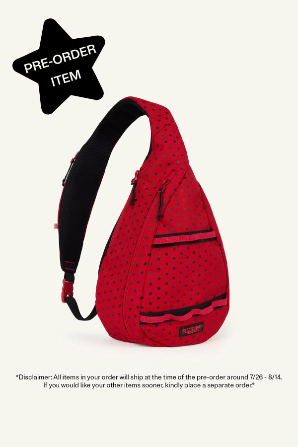 Product image of front view of a Red sling backpack with black polka dots, gray zippers, and a black adjustable strap. The bag has multiple front zip compartments. In the top left corner, a black star reads "Pre-Order Item." Bottom reads “Disclaimer: All items in your order will ship at the time of the pre-order around 7/26-8/14. If you would like your other items sooner, kindly place a separate order.