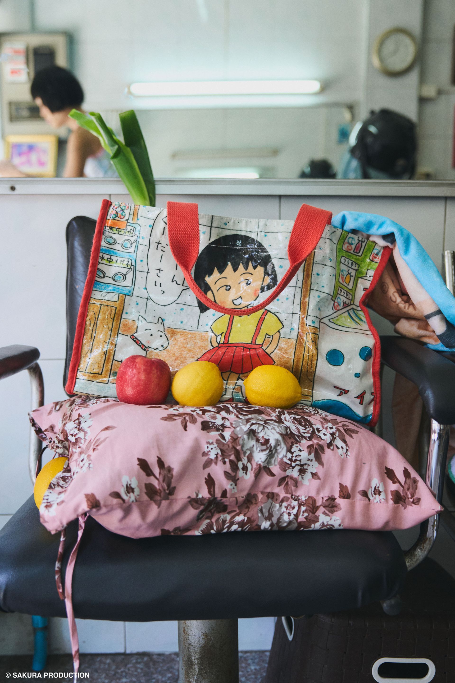 Carryall Bag sitting on top of a pillow with fruits. Shows image of manga drawing Chibi Maruko Chan and her dog walking into a store. She says "Grandma Please" in Japanese. Side panels red gingham print. Handles and trim are red. She wears the Sweet Shopper Tank, red gingham bikini skirt, and jeans.