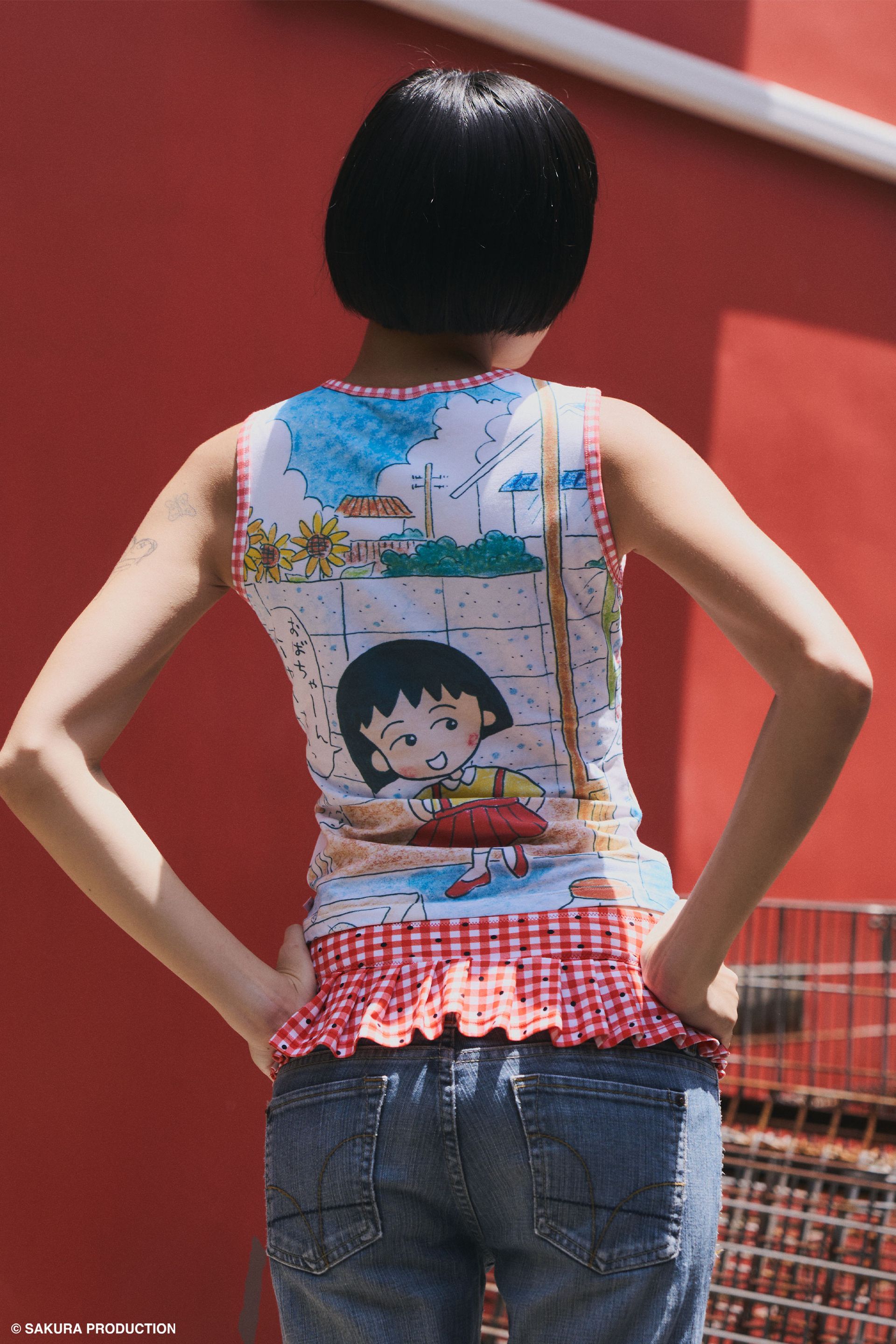 Back view of woman wearing Sweet Shopper Tank. Top illustrates a manga-style image of a young girl and her dog walking into a store. She speaks in Japanese, "Please Grandma!" The top has a red gingham trim around the neck and sleeves. She wears jeans and a red gingham swim bottom over them against a red background with crates.
