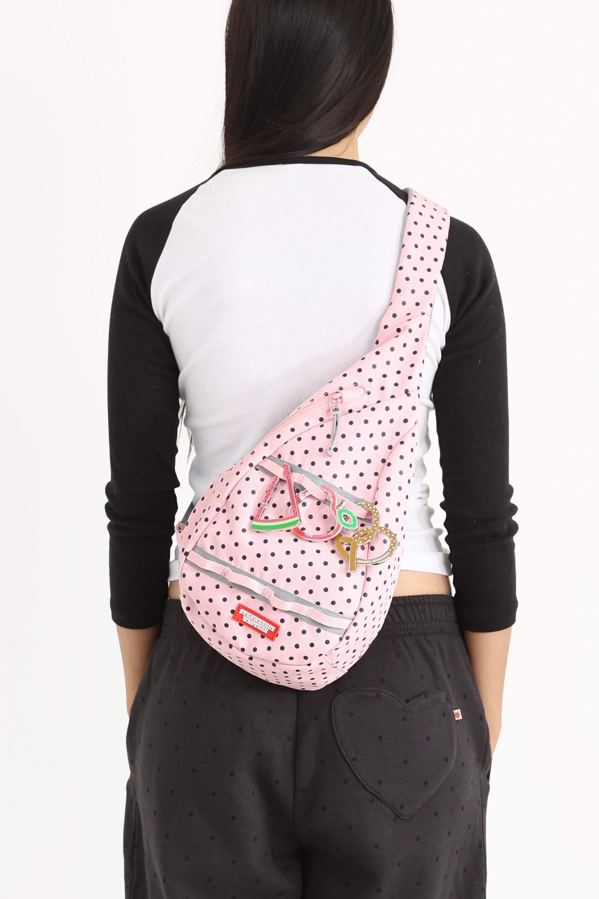 Back view of a model, She carries a pink polka-dotted sling bag across her back, decorated with colorful keychains shaped like fruits and food.