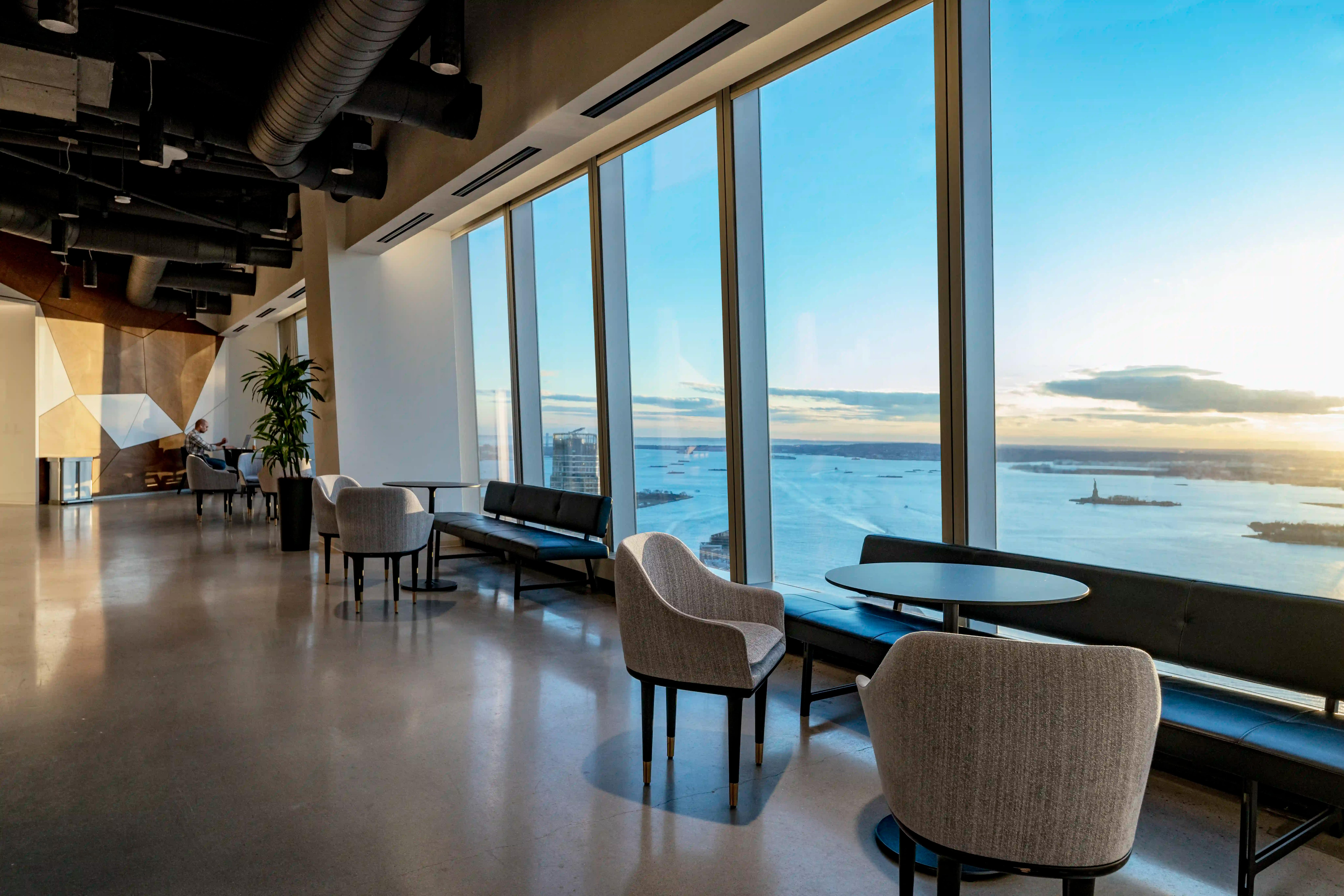 64th Floor of One World Trade Center. Modern office lounge with large windows offering a panoramic view of a city's waterfront at sunset, furnished with sofas and armchairs.