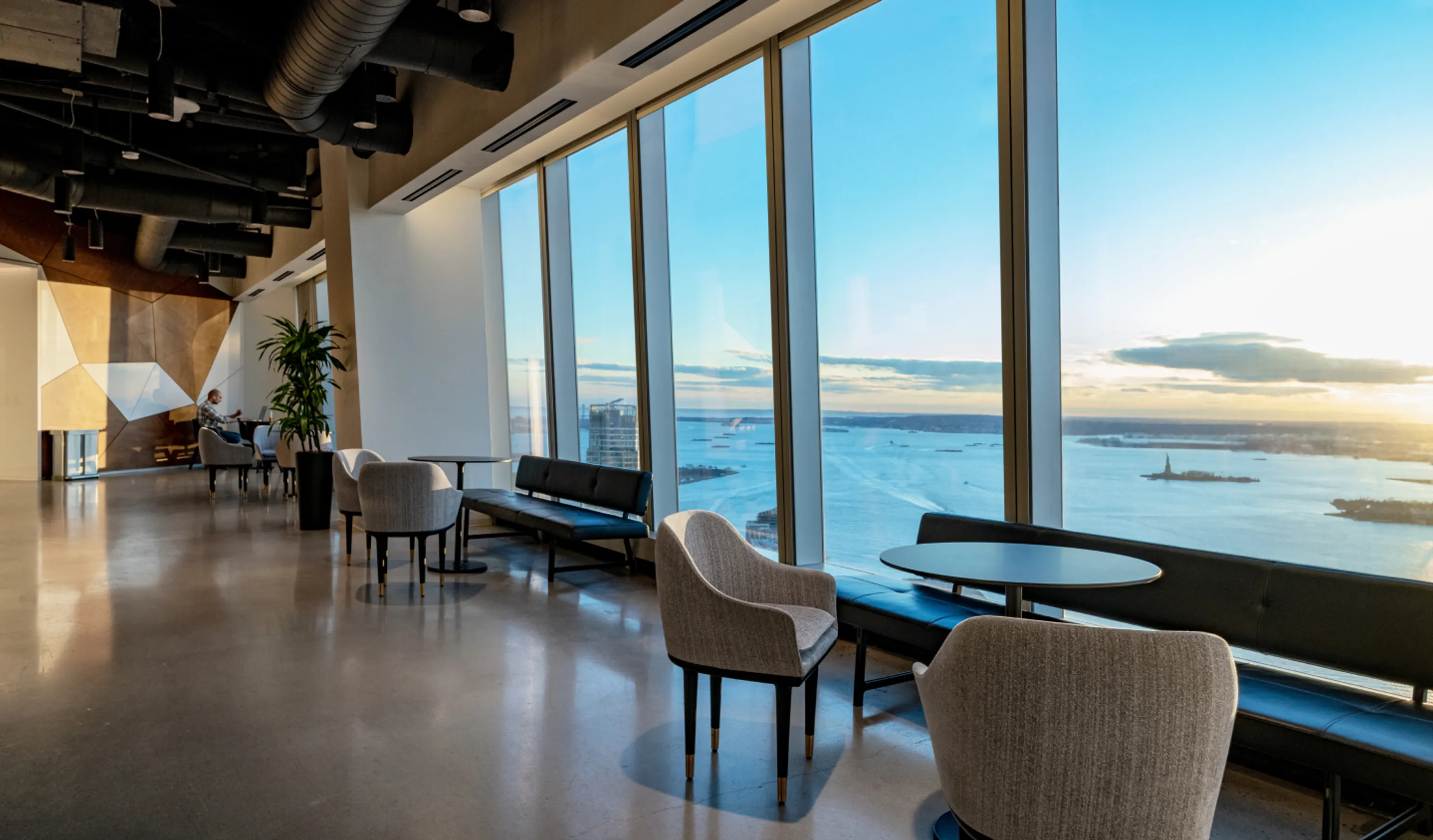 64th Floor of One World Trade Center. Modern office lounge with large windows offering a panoramic view of a city's waterfront at sunset, furnished with sofas and armchairs.