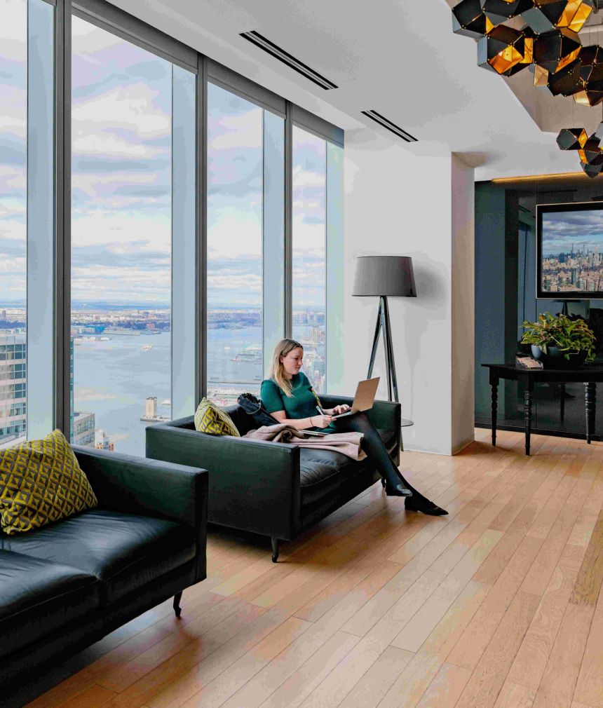 A woman sits on a couch using a laptop in a high-rise office with large windows overlooking the Hudson river and cityscape. Black couches and decorative pillows furnish the lounge area of One World Trade Center