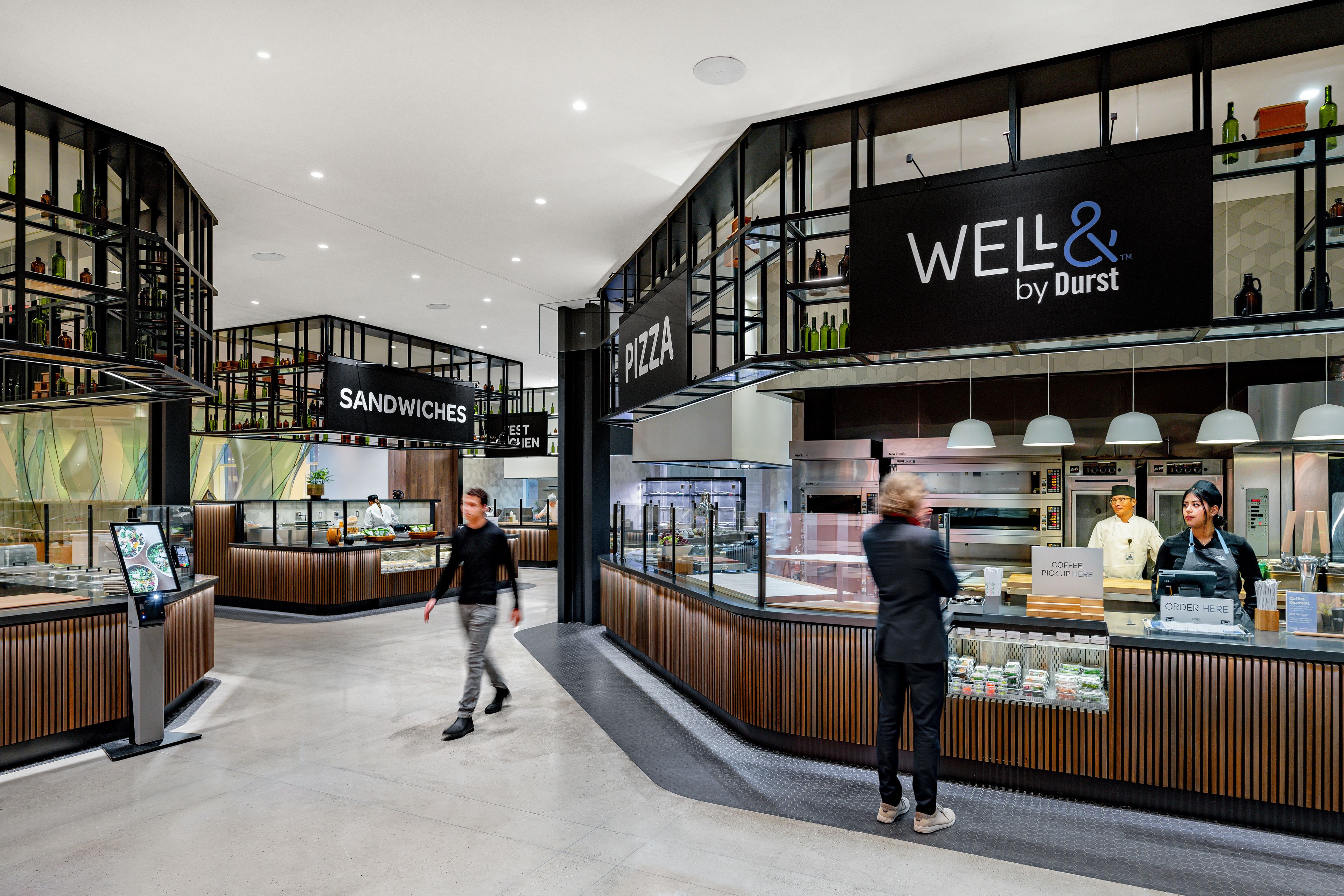 Modern cafeteria interior with a sign displaying "Well& by Durst", featuring sandwich and pizza stations, and seating area under soft lighting, with staff serving customers.