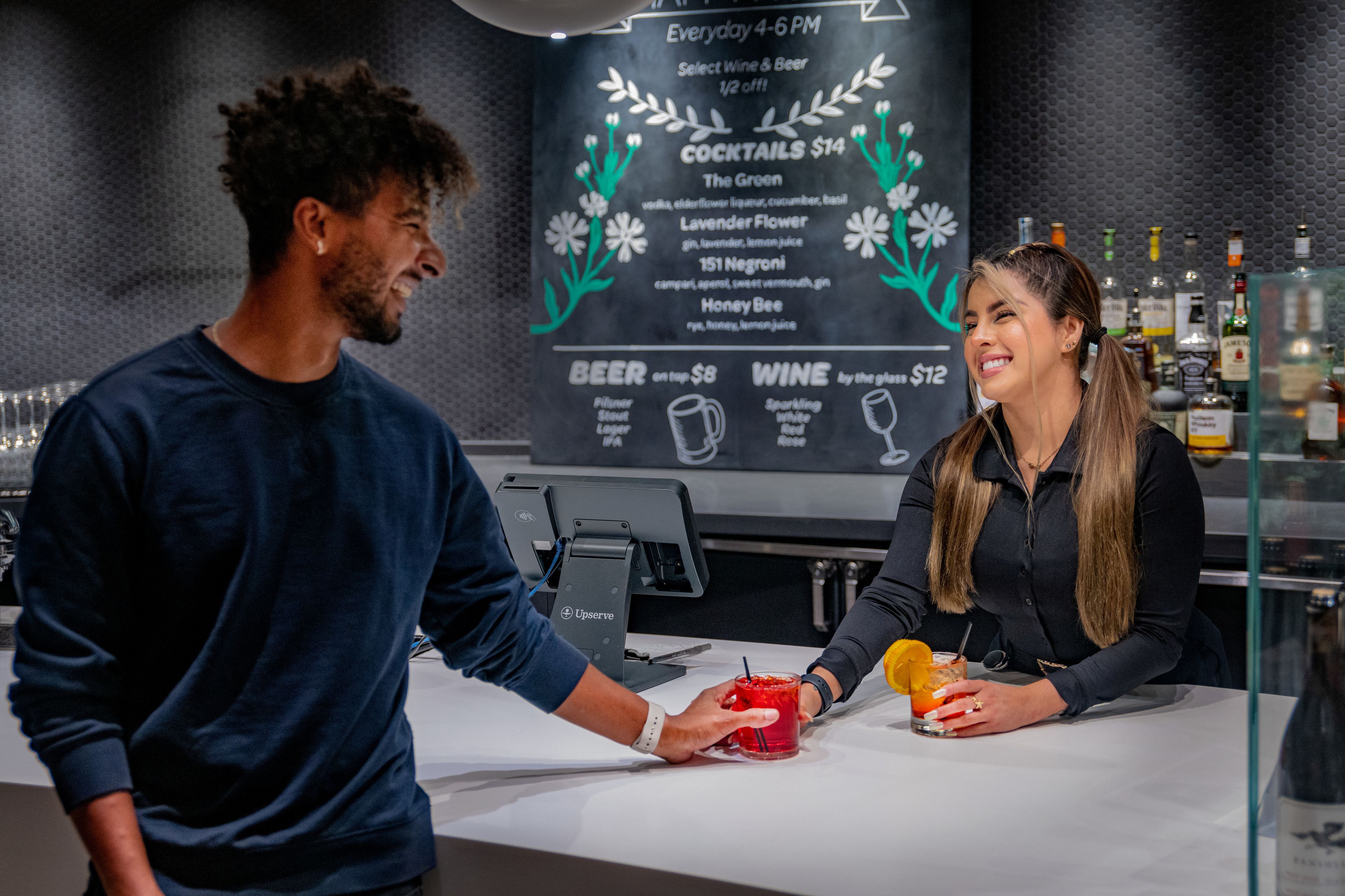Bartender serving a customer a cocktail at The Green with a chalkboard menu behind the bar.