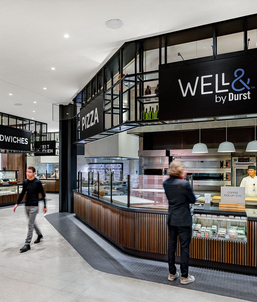 Modern cafeteria interior with a sign displaying "Well& by Durst", featuring sandwich and pizza stations, and seating area under soft lighting, with staff serving customers.