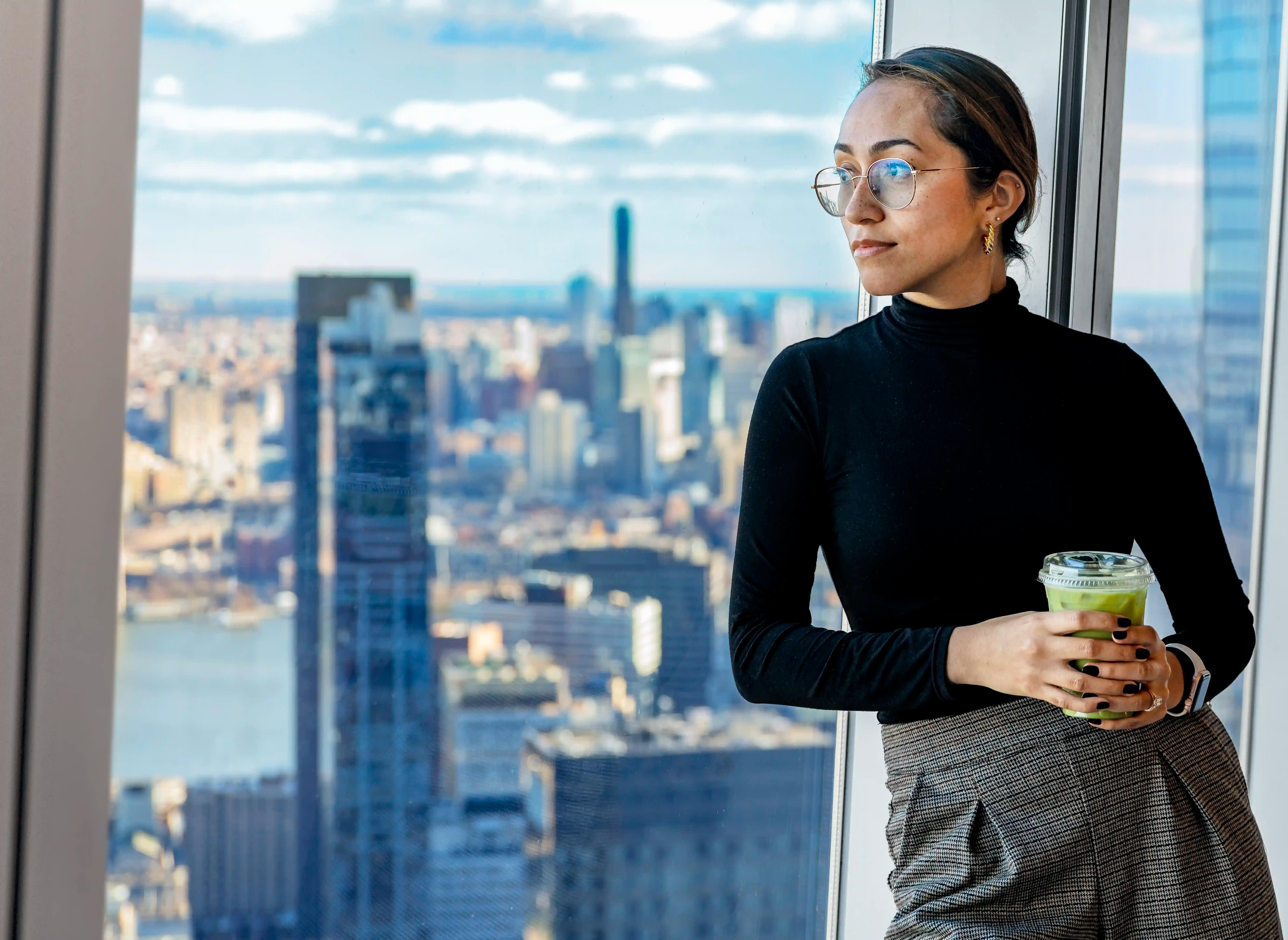 Woman wearing a black sweater, grey pants, glasses and with a cup with green juice in her hands leans against a window pane that overlooks the New York cityscape. This is from the SkyLobby on the 64th floor of One World Trade Center in their Well& by Durst cafe.