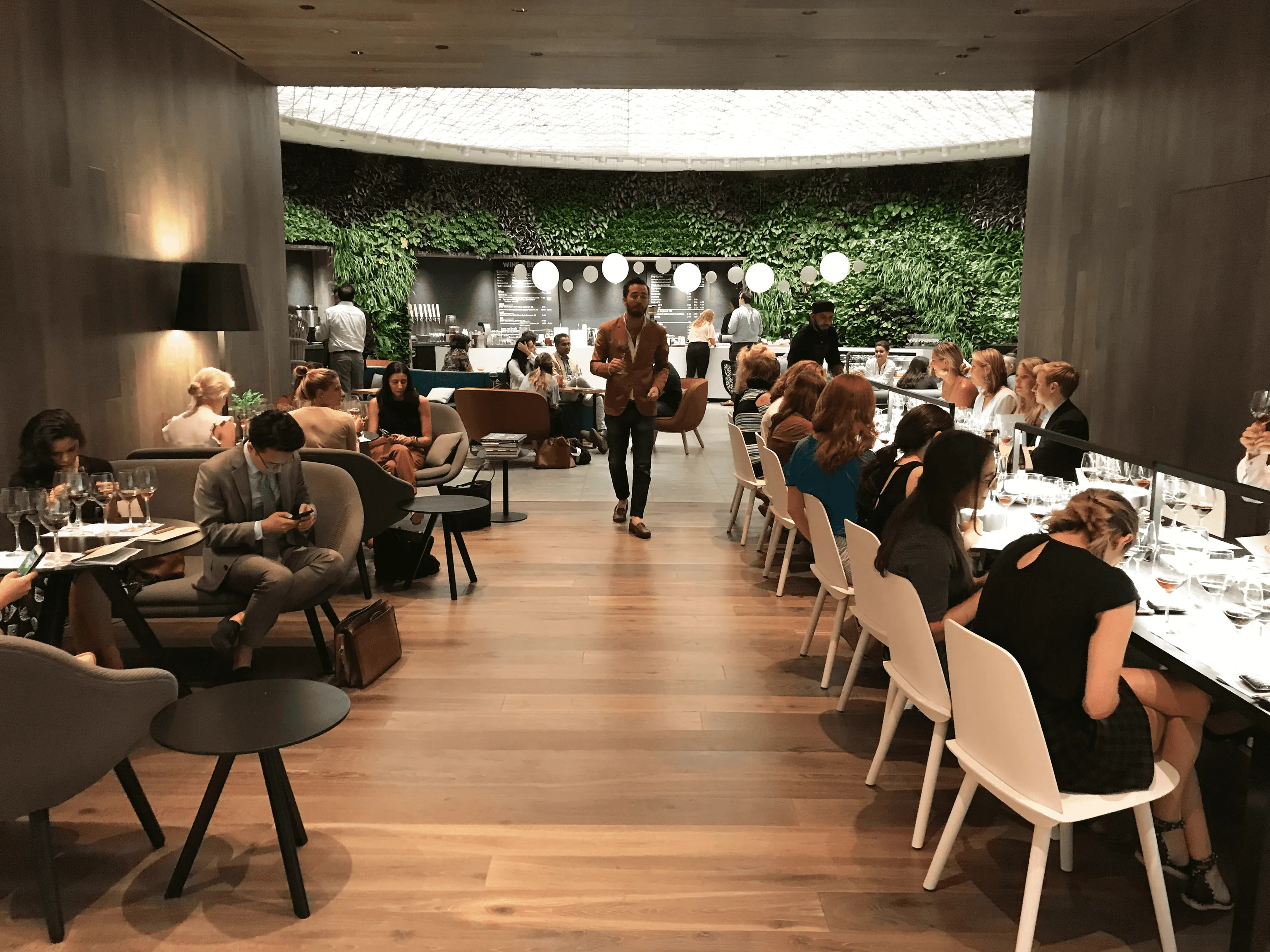 Bustling restaurant with a contemporary design, featuring a mix of long communal tables and smaller seating areas. Diners are engaged in conversation and enjoying their meals. A waiter is seen walking through the center of the room.