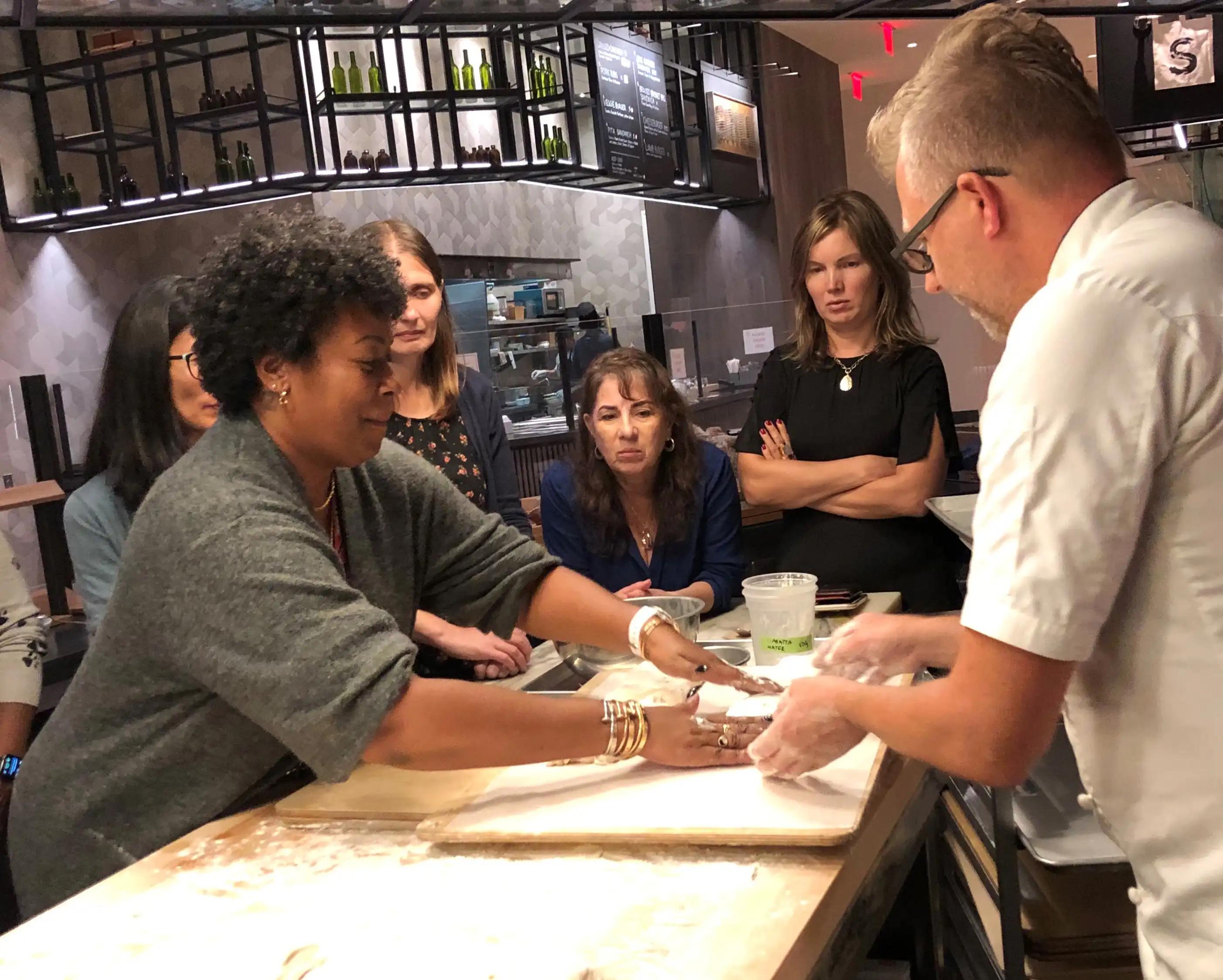 A woman kneading dough on a table surrounded by a group of people in a demonstration kitchen at Well& by Durst's 151 West 42nd street location.