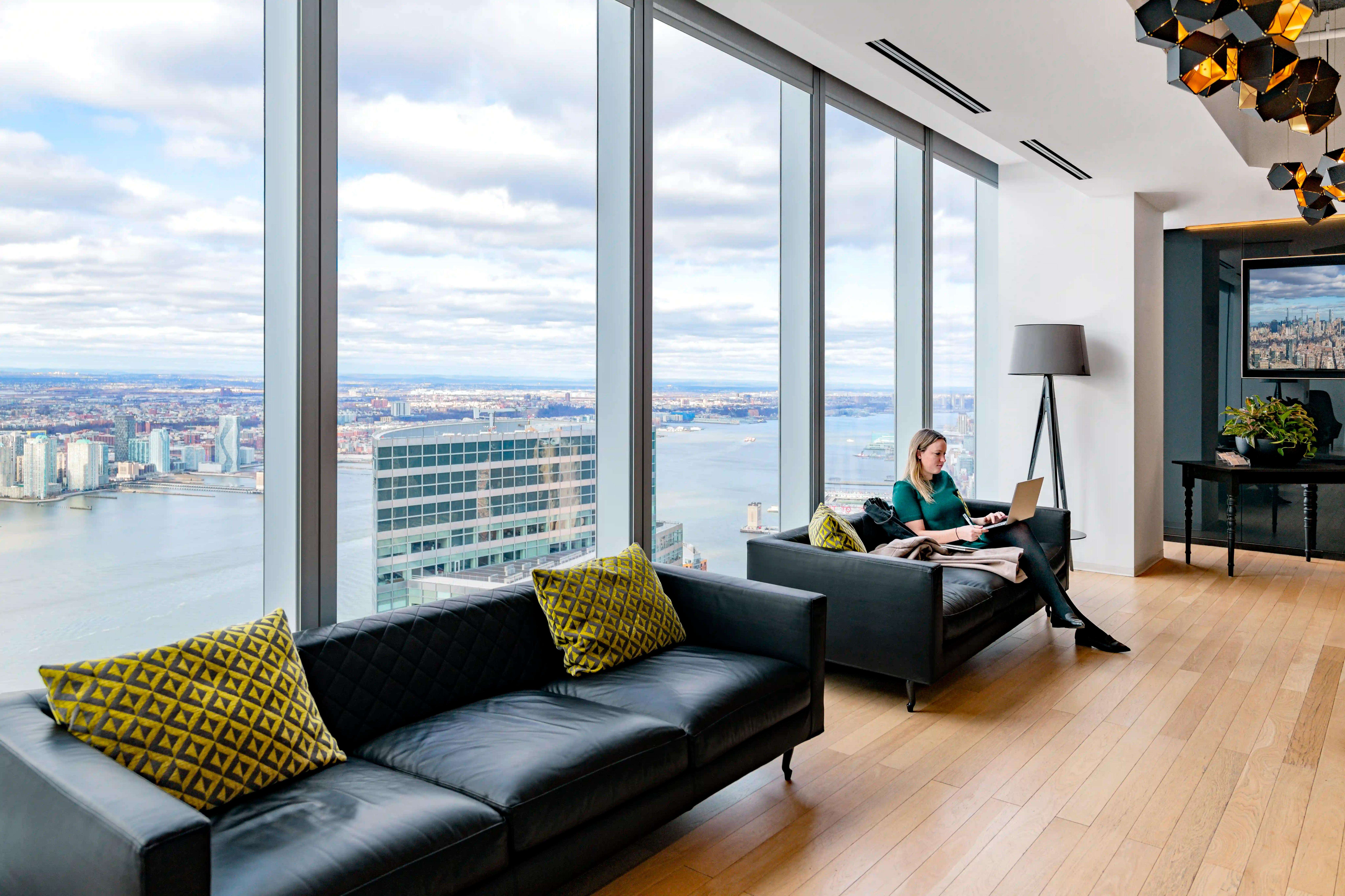 A woman sits on a couch using a laptop in a high-rise office with large windows overlooking the Hudson river and cityscape. Black couches and decorative pillows furnish the lounge area of One World Trade Center