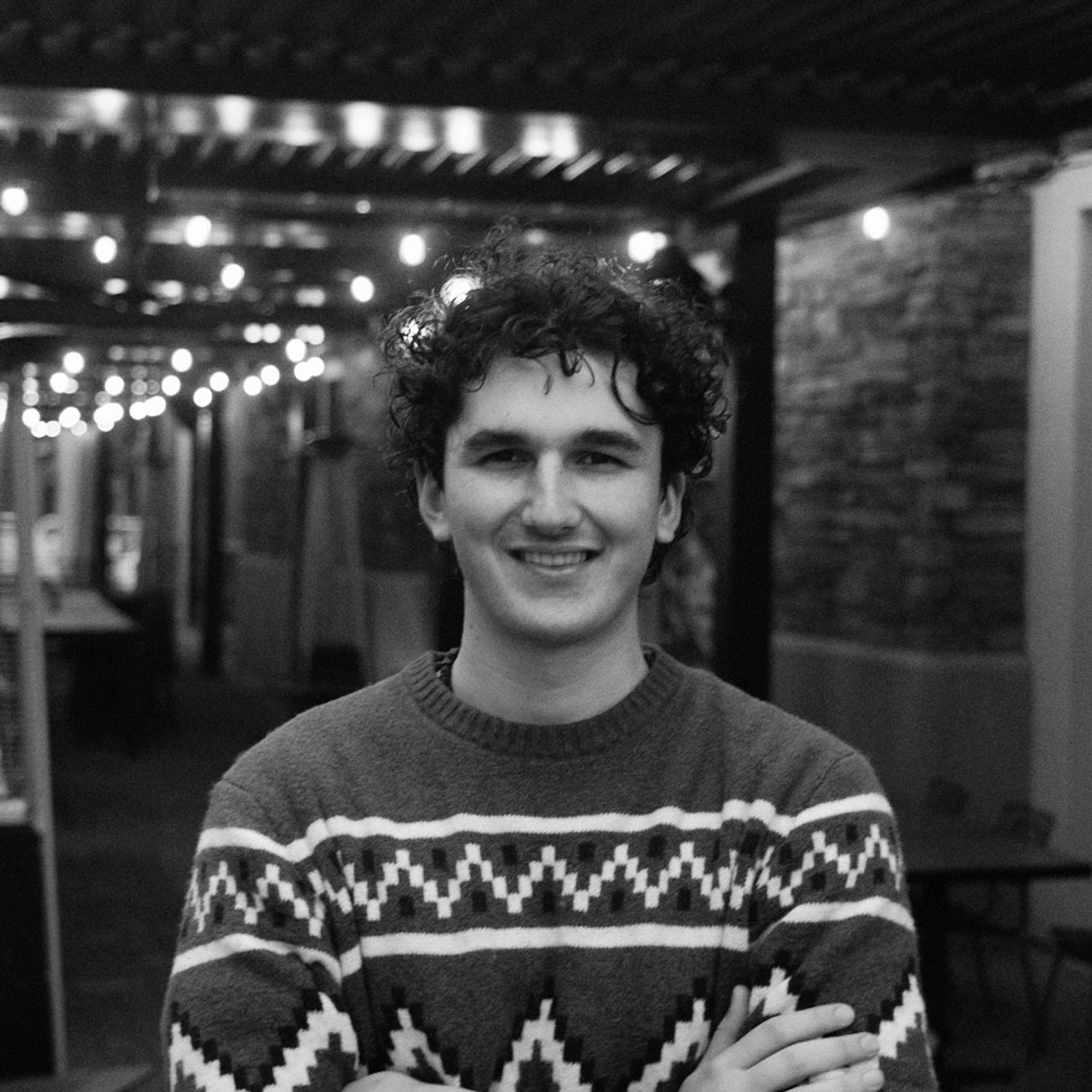 Joe Le Flore, black and white image, standing in a brick and concrete alley with cafe lighting and a decorative sweater