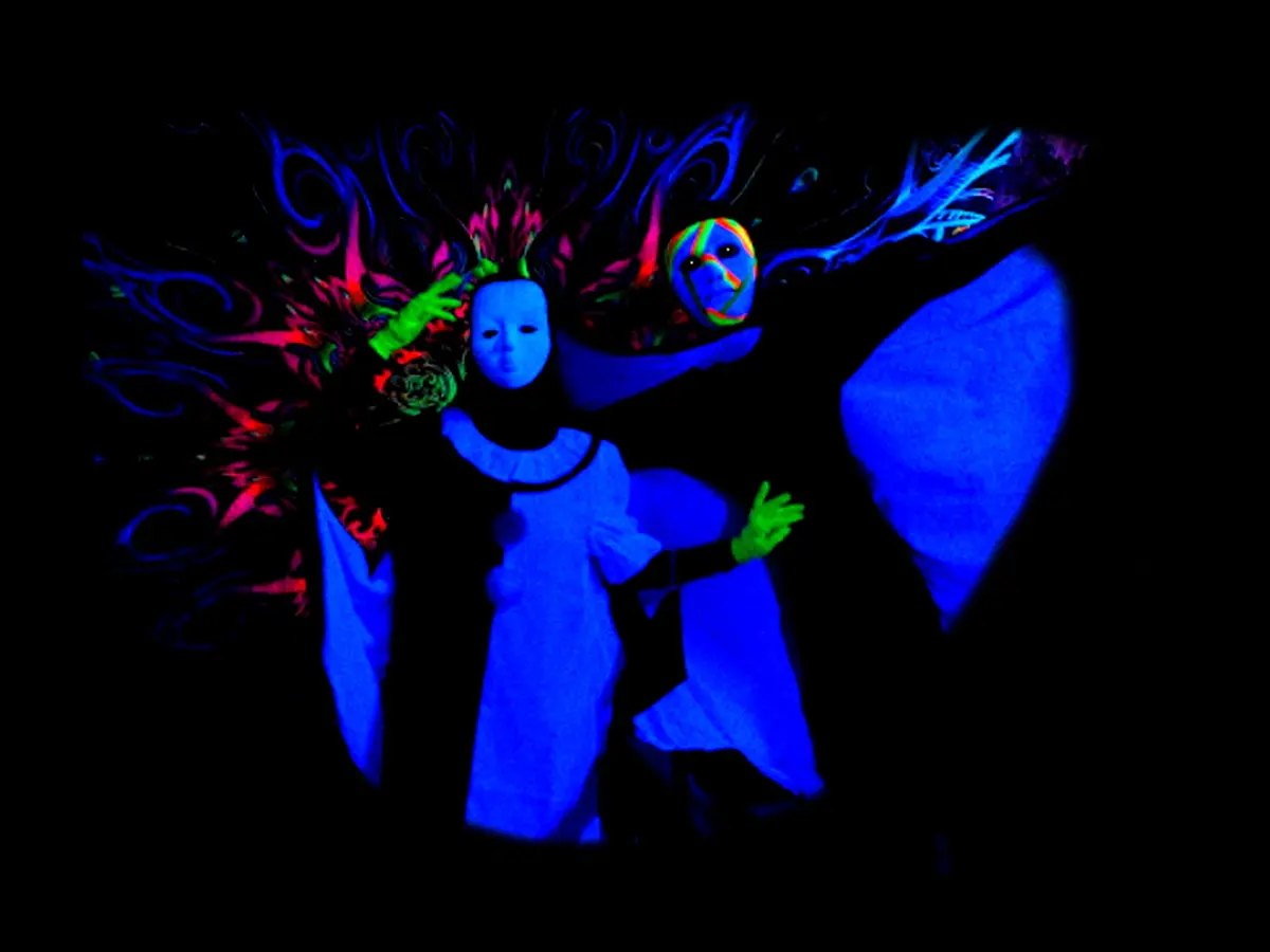 Escena de teatro negro con figuras y máscaras fluorescentes bajo luz ultravioleta