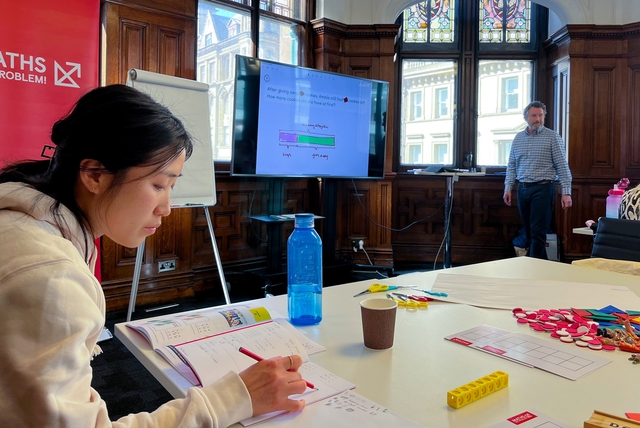 Asian woman at Maths — No Problem! training event with Maths — No Problem! textbooks in front of her and a notebook to take notes in. Adam Gifford in the background next to presentation.