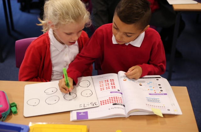 A girl and a boy working on maths together with a whiteboard and a textbook in front of them.