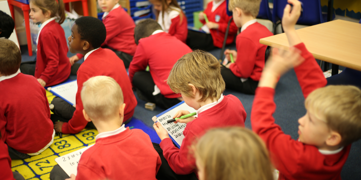 Young children with whiteboards, writing down their answers. One boy raises his hand to ask a question.
