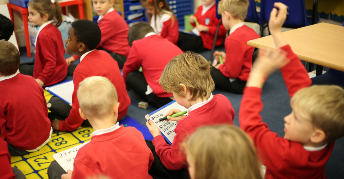 Young children with whiteboards, writing down their answers. One boy raises his hand to ask a question.