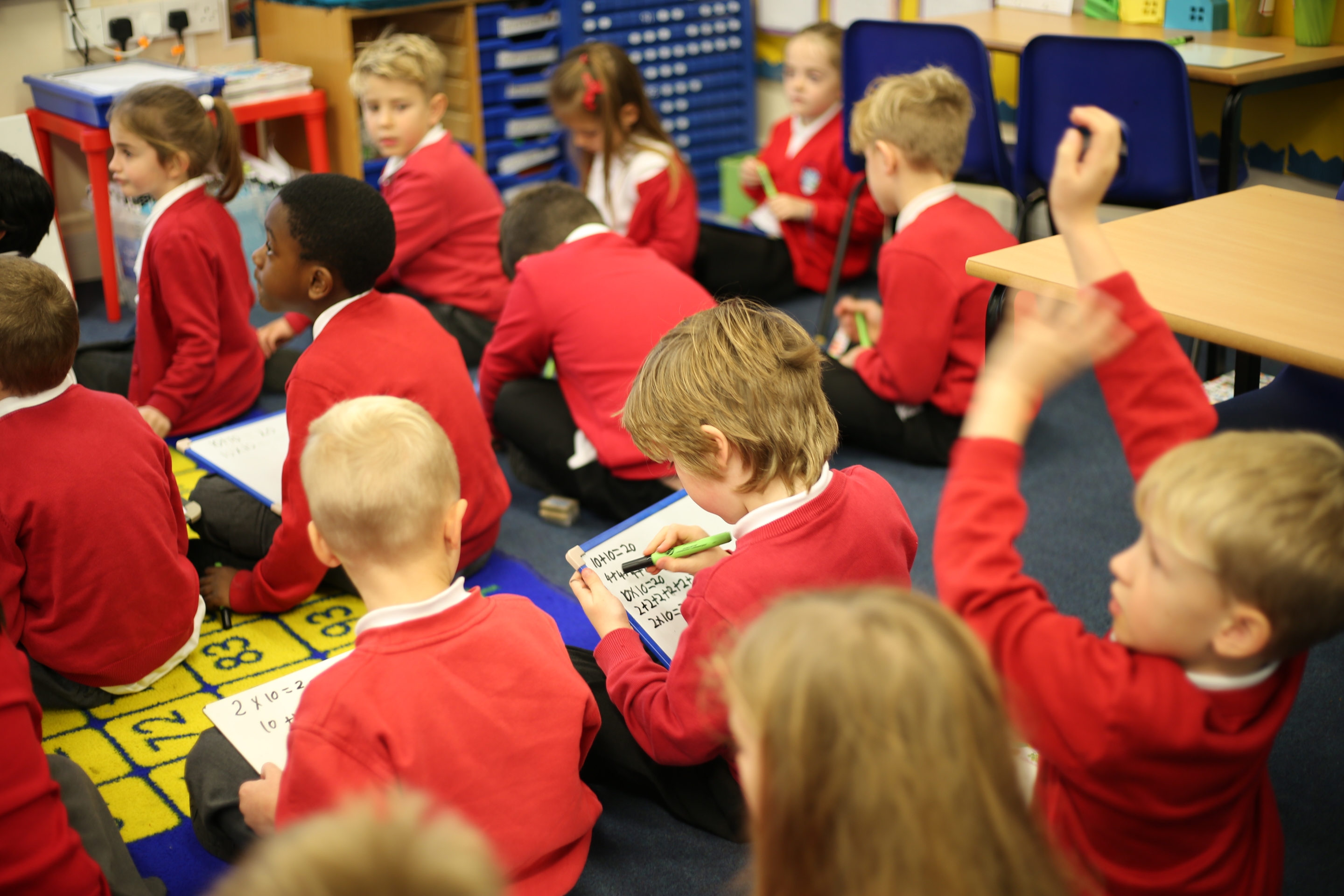 A diverse group of pupils in red uniforms sitting on the floor of a colourful classroom, working on math problems