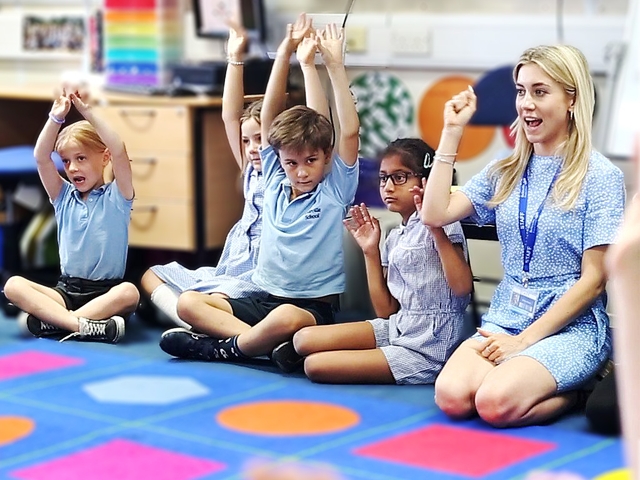 Teacher teaching on the floor with young pupils who are raising their hands.