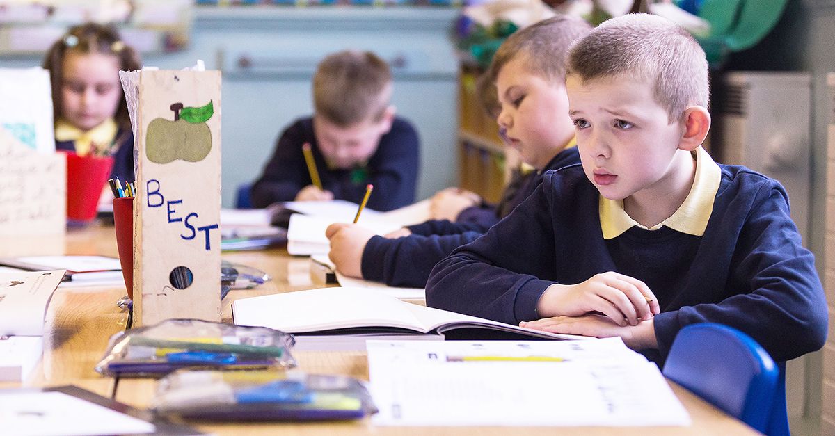 One male student with a confused look on his face surrounded by fellow pupils who are working in their workbooks.
