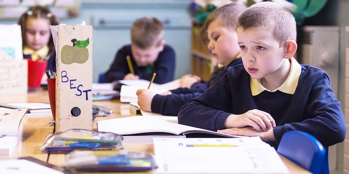 One male student with a confused look on his face surrounded by fellow pupils who are working in their workbooks.