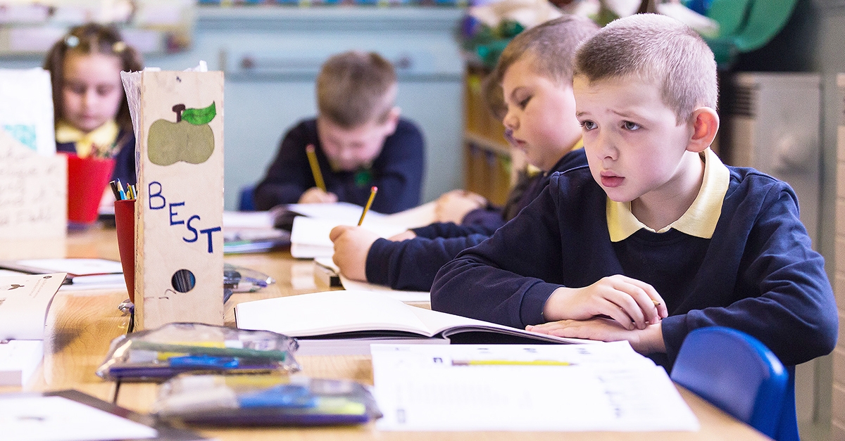 One male student with a confused look on his face surrounded by fellow pupils who are working in their workbooks.