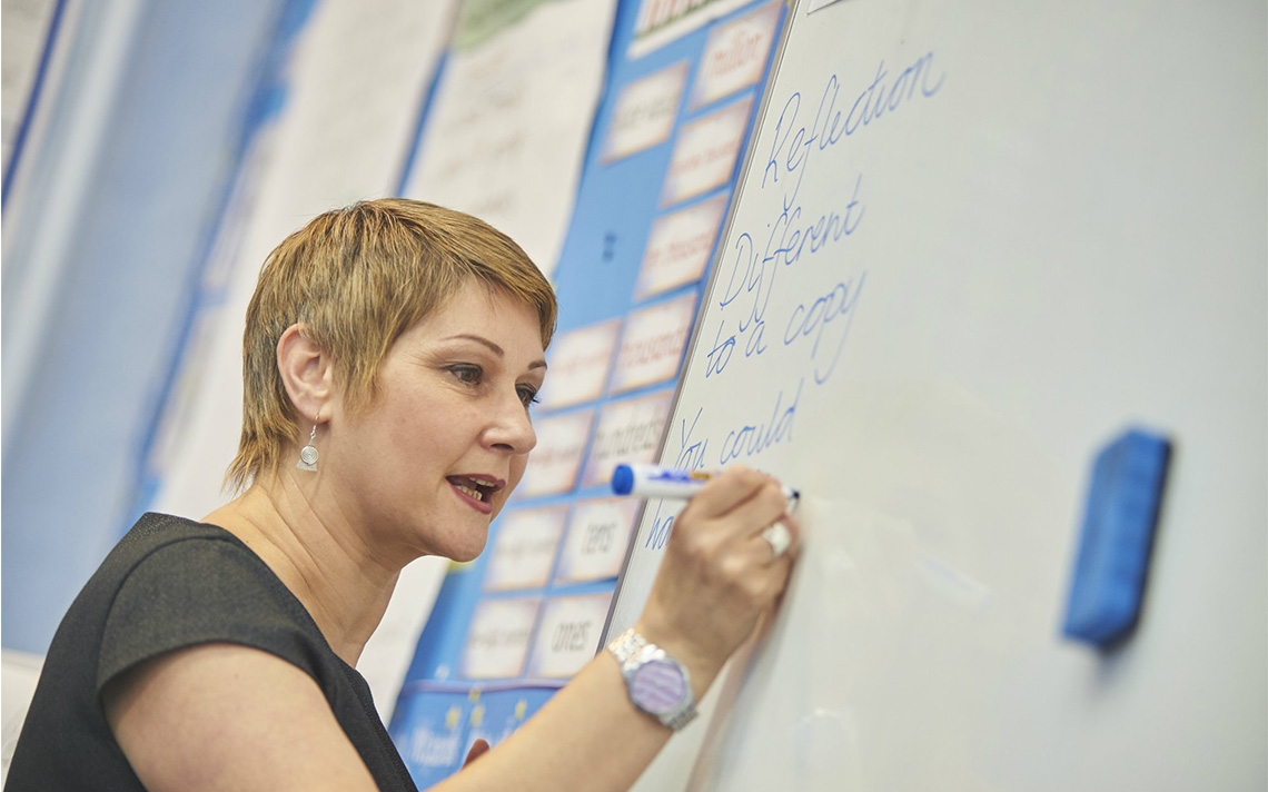 A maths teacher writing on a whiteboard in blue marker