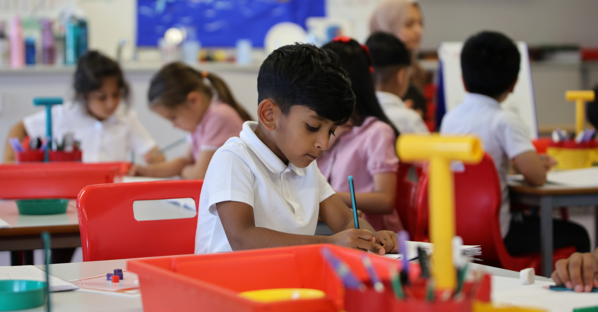 A young male pupil writing at his desk. Other pupils are in the backhro