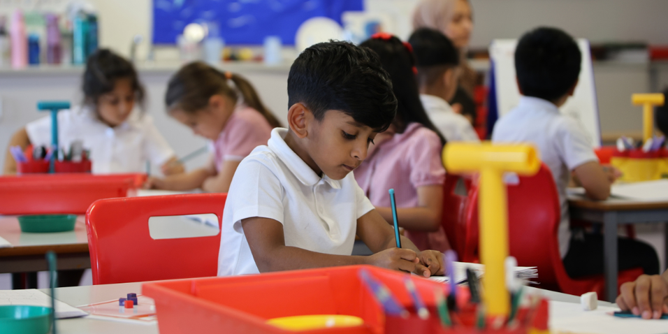 A young male pupil writing at his desk. Other pupils are in the backhro