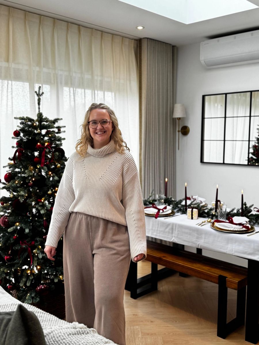 A smiling woman wearing glasses, a beige ribbed jumper and wide-leg trousers stands in a living room decorated for Christmas, with a decorated Christmas tree and festive dining table in the background.