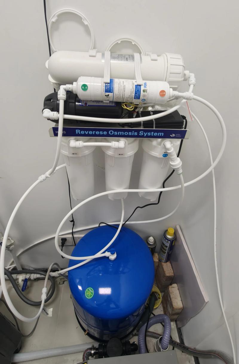 A wall-mounted 5-stage Reverse Osmosis (RO) water filtration system with three white vertical filter canisters (PP, UDF, CTO) and a horizontal RO membrane housing. Below it sits a large blue pressurized water storage tank connected by white tubing.