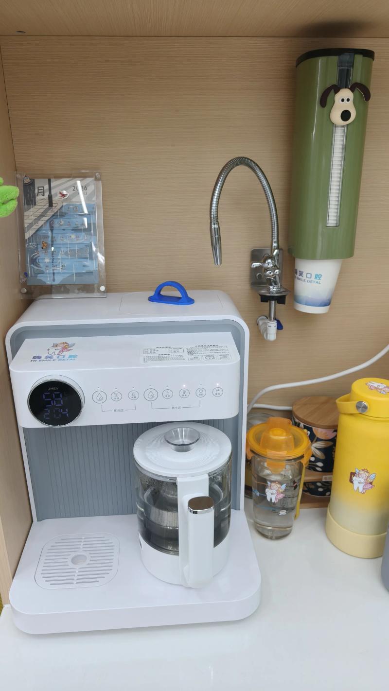 A white countertop smart water dispenser with a glass pitcher and digital display, located in a dental clinic. Next to it is a wall-mounted faucet and a green cup dispenser. The equipment is decorated with "Hi Smile Dental" branding stickers.