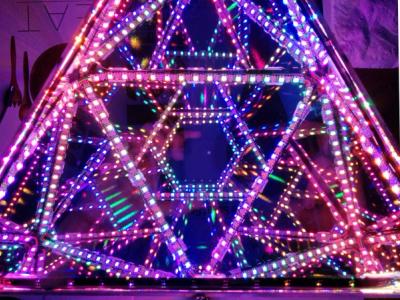 An octahedral infinity mirror with repeating patterns of glowing LEDs, reflecting internally in pinks, purples, and greens