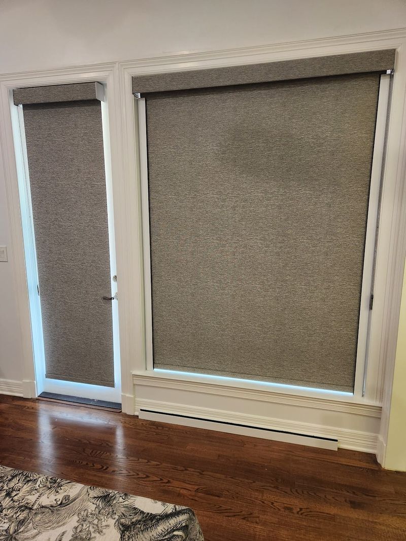 Gray roller shades fully lowered on a large window and a narrow door in a bright room with hardwood floors