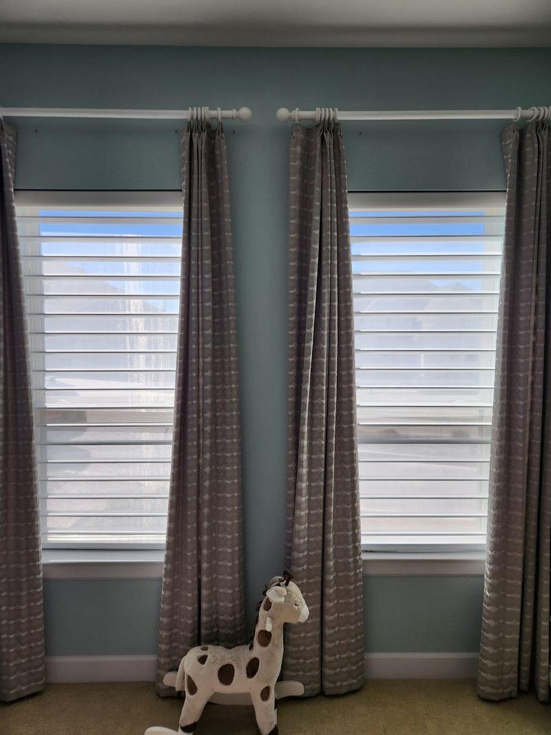 Gray patterned drapery panels with sheer horizontal shades on two windows in a nursery