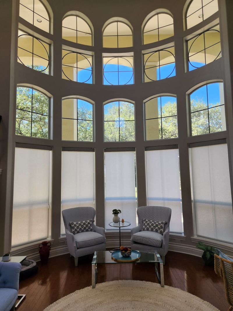 White solar roller shades on tall arched windows in a two-story living room with two accent chairs