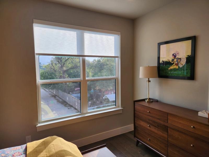 Manual roller shade partially lowered on large double window in modern bedroom with wood dresser