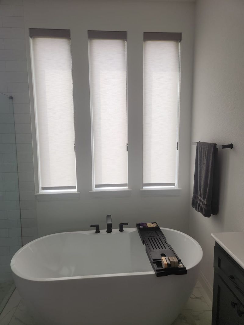 Three white cellular shades on tall narrow windows above a freestanding soaking tub in a modern bathroom