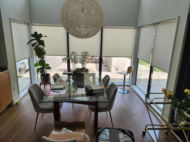 Cream roller shades on floor-to-ceiling windows in modern dining room with glass table