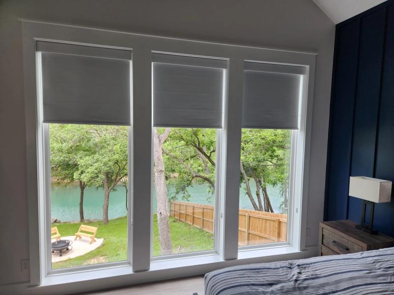 Three gray roller shades partially lowered on large bedroom windows overlooking a lake