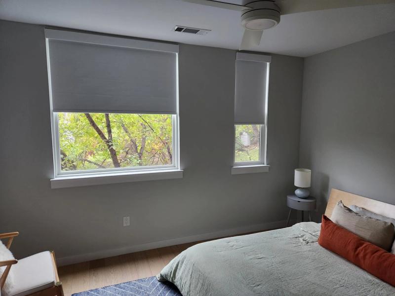 Gray roller shades partially lowered on two bedroom windows with green tree view