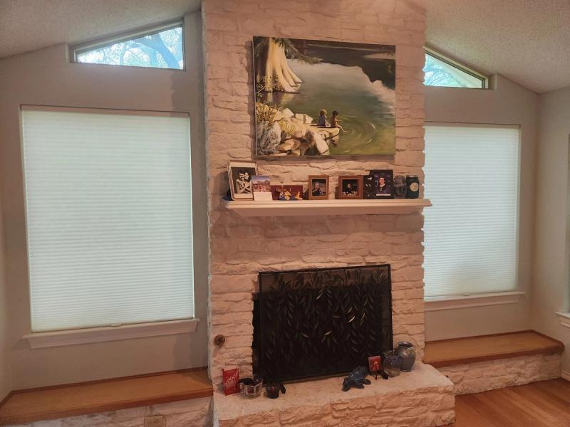 White cellular honeycomb shades on two living room windows flanking a painted stone fireplace