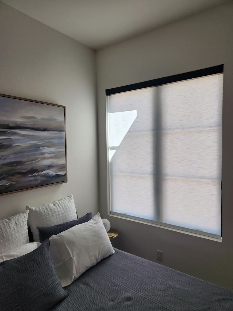 Solar roller shades on a large bedroom window beside a bed with gray bedding